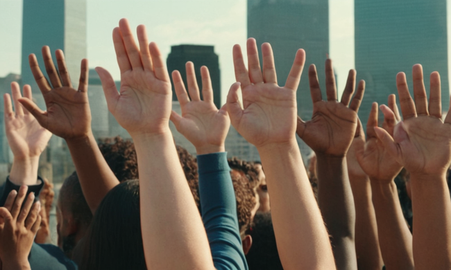 Grupo de personas de diversos orígenes que se unen con las manos levantadas en solidaridad, frente a un paisaje urbano.