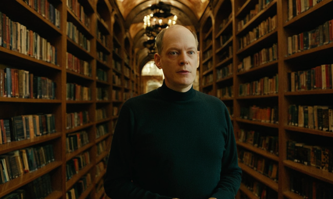 Alain de Botton de pie en medio de citas filosóficas en estanterías rodeadas de retratos pensadores.