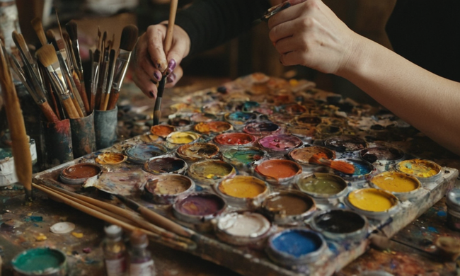 Las manos de los artistas trabajando en un lienzo con pinceles dispersos y tubos de pintura en el fondo.